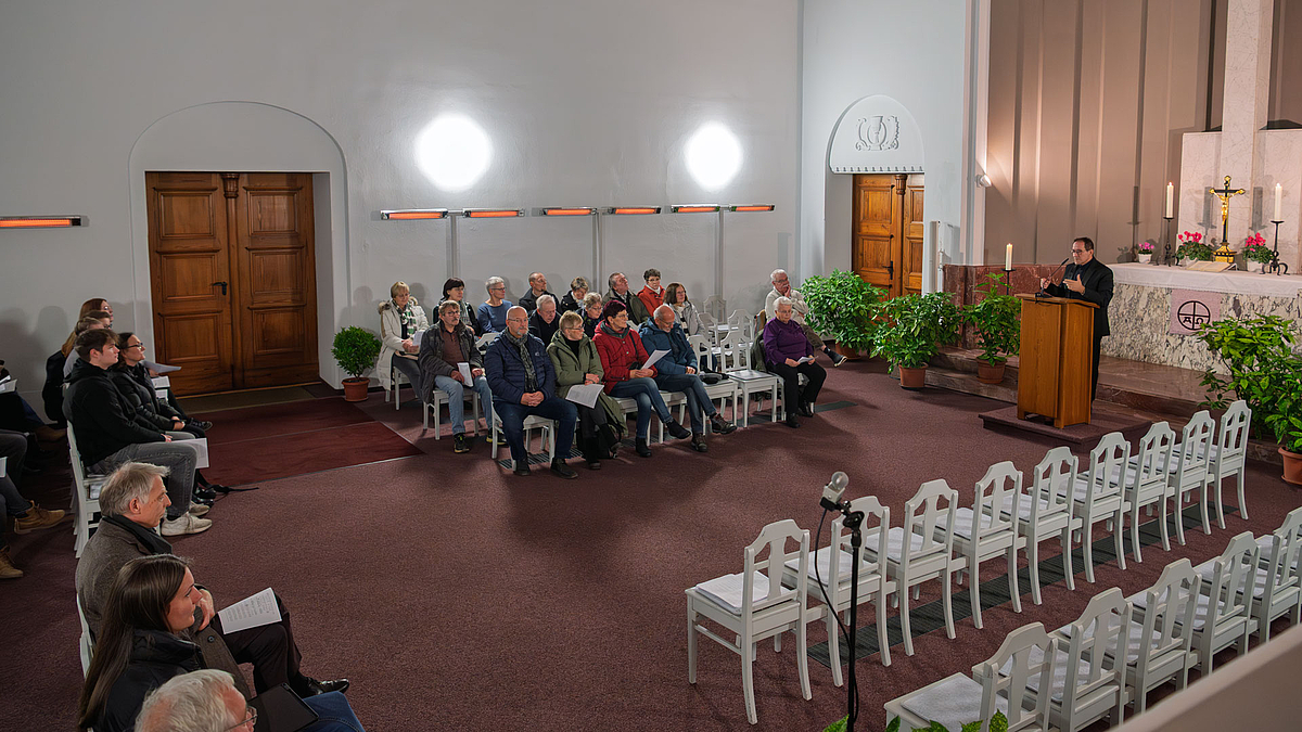 Blick in den großen Kapellenraum mit Besuchenden auf wei0en Stühlen. Am Rdnerpult vor inen Aktarr mit Kerzen und einem großen Kreuz eine männliche Person im Anzug.