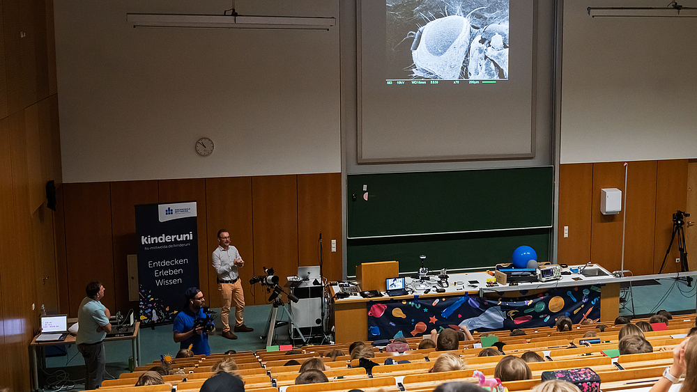Blick von oben auf den Hösraal mit Kindern udn Dozenten. Auf der Leinwand ist die starke Vergrößerung der Augen einer Fruchtfliege zu sehen.