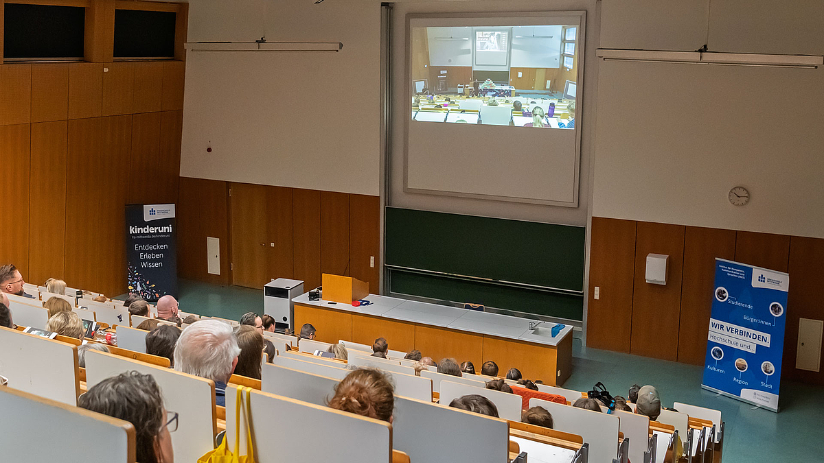In einem Hörsaal mit vielen Sitzreihen sitzen Menschen, die nach vorn auf eine Leinwand schauen. Auf der Leinwand ist das Geschehen der Kinderuni zu sehen, das übertragen wird.