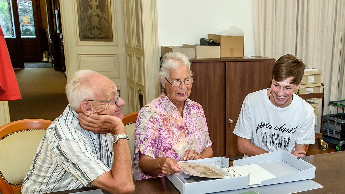 Das Foto zeigt das ältere Ehepaar Knieper mit ihrem Enkel an einem großen Tisch im Hochschularchiv. Sie blättern interessiert und gut gelaunt in einem Archivordner mit alten Fotografien.