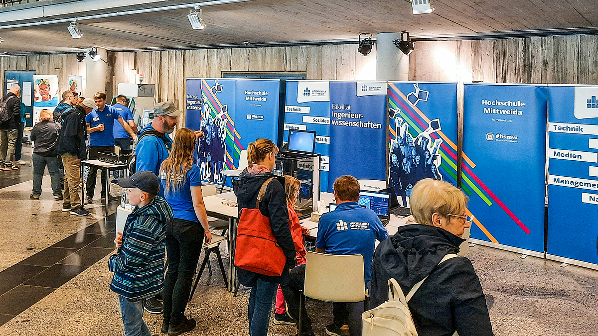 Ein breiter Messestand in einer Halle. Im Hintergrund stehen mehrere blaue Rollups der Hochschule Mittweida, darunter in der Mitte eines mit der Aufschrift „Fakultät Ingenieurwissenschaften“. Davor stehen Tische mit Geräten. Vier Standbetreuende mit „Hochschule Mittweida“-Poloshirt sowie zahlreiche Besuchende stehen oder sitzen an den Tischen.