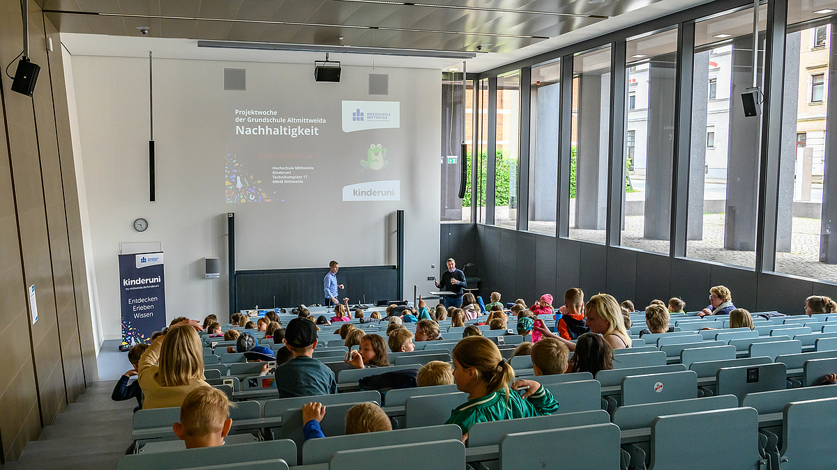 Blick über die Köpfe der Kinder hinweg auf die beiden Dozenten am unteren Ende des Hörsaals. Dort steht links ein Rollup mit dem Logo der Hochschule und dem Text „kinderuni. Entdecken. Erleben. Wissen“. An die Rückwand ist eine Folie projiziert mit dem Titel der Vorlesung „Projektwoche der Grundschule Altmittweida. Nachhaltigkeit.“