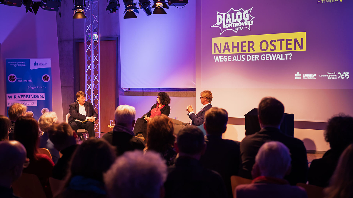 Blicl über die Köpfe von Zuschauenden hinweg auf drei Personen des Podiusm. Über ihnen auf der Leinwand ist "Dialog Kontrovers Extra. Naher Osten. Wege aus der Gewalt" zu lesen".