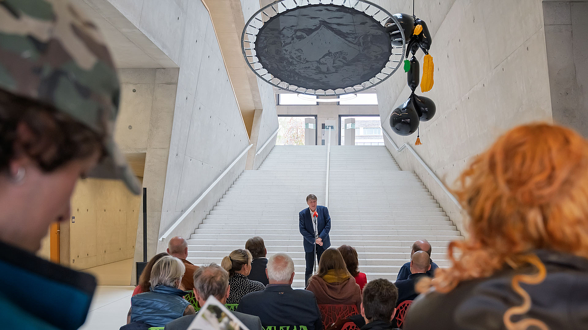 Biick durch Zuschauende jidurch auf eine große Treppe, an deren  Fuß eine männliche Person im Anzug steht und spricht. Über ihn hängenn überdimensionale Früchte und ein überdimensionales Trampolin, auf dessen Matte u.a. ein Zelt gemalt ist.