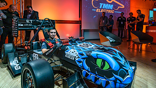 Probesitzen im neuen Auto: TMM-Technikchef Mika Schuster. Mit Black Mamba startet das Motorsport-Team der Hochschule Mittweida in die neue Formula-Student-Saison. Das Foto zeigt einen jungen Mann im Rennwagen sitzend. Der Rennwagen befindet sich in einem großen Raum. Hinter dem Rennwagen stehen Personen, die Details begutachten. Im Hintergrund sind weitere Personen zu sehen, die das Auto bestaunen.