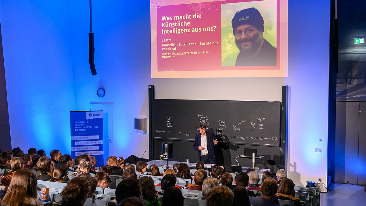 Blick von hinten über das Publikum im Hörsaal. Vor der Tafel steht eine männliche Person, die zum Publikum spricht. Auf dem über ihr projizierten Beamerbild ist rechts neben dem Porträt einer männlichen Person zu lesen: „Was macht die Künstliche Intelligenz aus uns? 9.5.2023. Künstliche Intelligenz – Büchse der Pandora? Prof. Dr. Thomas Villmann, Hochschule Mittweida“.