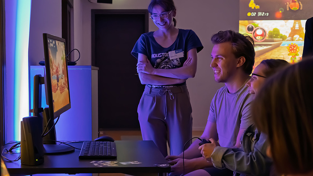 Seiitlicher Blick auf zwei junge Personen, die begeistert ein Computerspiel spielen. Eine junge weibliche stehende Person schaut ebenso begeistert zu.