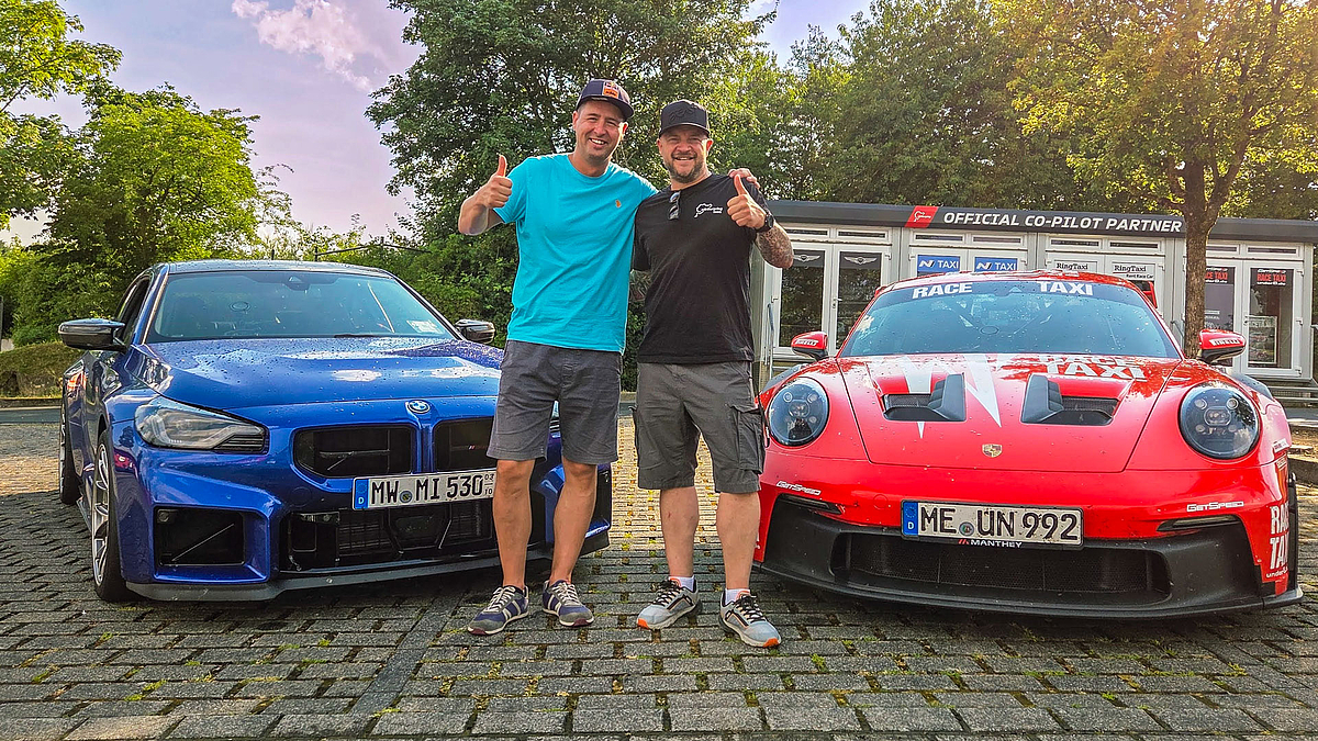 Prof und Profi: Professor Matthias Vodel und Rennfahrer Andreas Gülden. (Foto: privat) Zwei männliche Personen stehen die Arme gegebsitig um die Schultern zwischen ihren Renwagen, einem blauem BMW M2 und einem roten Porsche 911 (992)