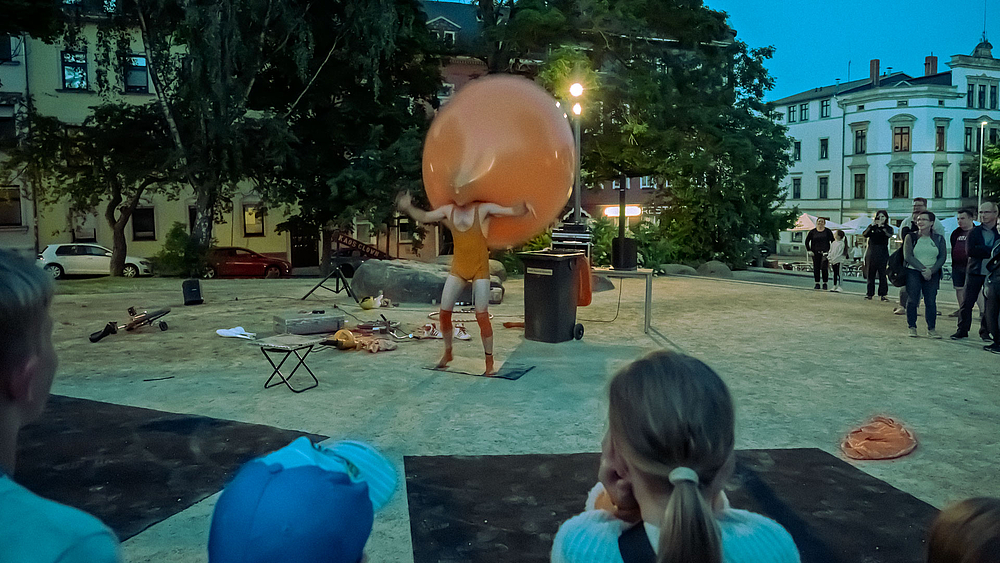 Der Clown steht mit sienem Kopf in einem riesigen orangenen Ballon auf dem Platz.