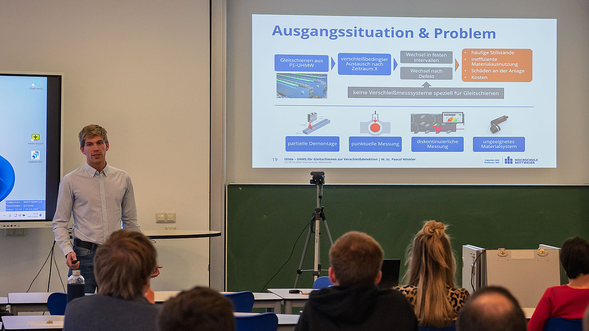 Männliche Person vor projizierter Powerpointpräsentation. Im Vordergrund Köpfe des Publikums von hinten.