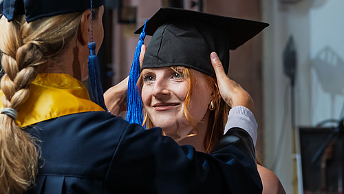 Weibliche Person im Talar und mit Doktorhut setzt Absolventin den Graduierten-Hut auf.