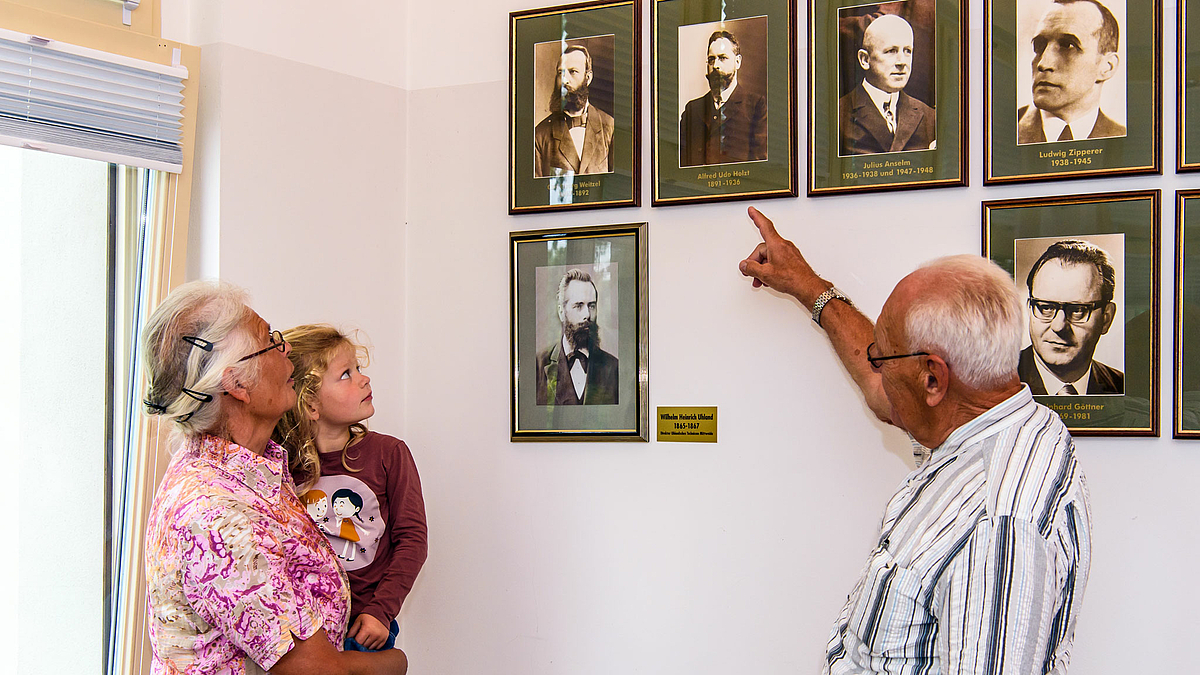 Das Foto zeigt eine ältere Frau mit ihrer vierjährigen Enkelin auf dem Arm. Gemeinsam mit einem älteren Mann stehen Sie vor der Bildergalerie mit den Porträts der früheren Direktoren der Hochschule Mittweida. Der ältere Mann ist Axel Knieper, der Urenkel von Alfred Udo Holzt. Er weist mit dem Finger auf des Porträt Holzts an der Wand und zeigt seiner Frau und seiner Enkelin den Vorfahren.