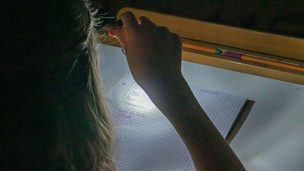 Ein Kind sitzt im Dunkeln und beleuchtet mit einer Taschenlampe ein kariertes Heft mit eigenen Notizen.