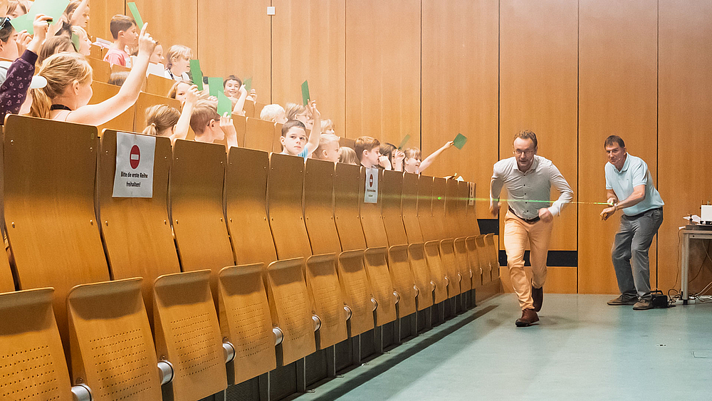 Ein Dozent erzeugt einen Lichtsrahl am Fuß des Hörsaals. Sein Kollege versucht mit einem Sprint schneller am anderen Ende anzukommen als das Licht.