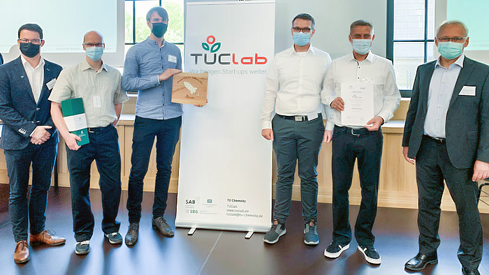 Das Fotos zeigt sechs männliche Personen mit Corona-Maske links und rechst von einem Rollup mit der Aufschrift "TUClab". Drei der Personen halten Urkunden und ein Geschenk. 