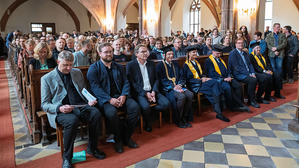 Blick ins vollbesetzte Kirchenschiff. Die erste Bankreihe mit Akteur:innen, teilweise in bauen und schwarzen Talaren und mit Doktorhüten.