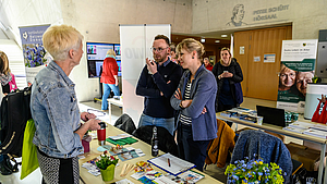 Auf Tischen im Foyer liegen zahlreiche Broschüren. Hinter einem der Tische stehen eine weibliche und eine männliche Person, die sich mit einer weiblichen Person vor dem Tisch unterhalten. Im Hintergrund sind Rollups und weitere Menschen zu sehen.