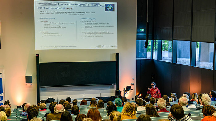 Blick von hinten in den Hörsaal über die Köpfe des Publikums auf den männlichen Dozenten. Schräg über ihm ist eine Folie projiziert mit der Überschrift: „Anwendungen von KI und maschinellem Lernen – III – ChatGPT & Co“ und „Was ist, was kann ChatGPT ¬ nicht?“