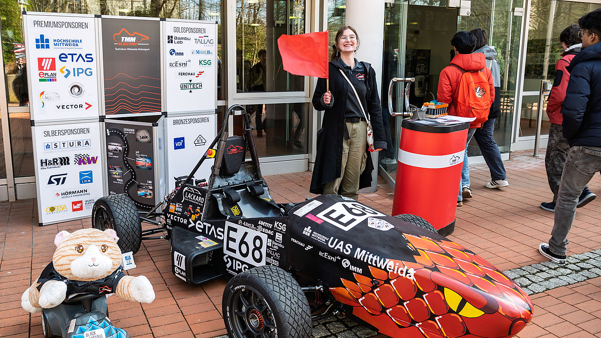 Junge weibliche OPerson steht hinter einem Rennwagen, schwenkt eine rote Flagge. Hinter ihr strömen Menschen in ein Gebäude.