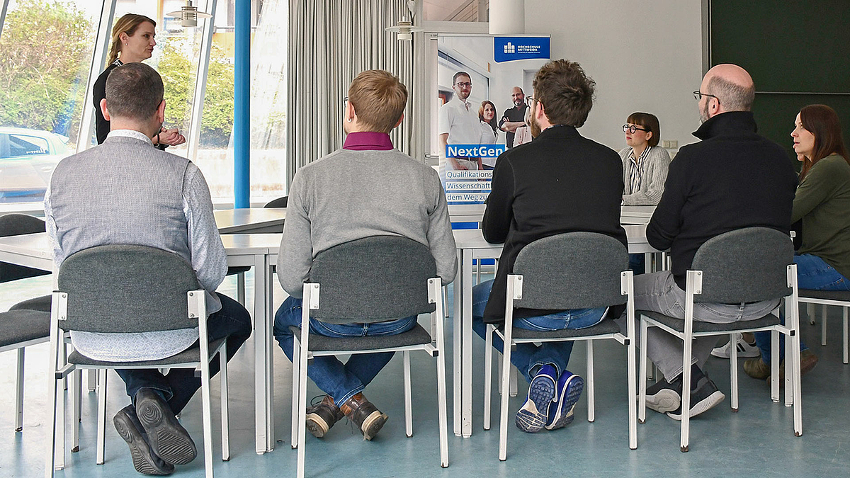 Im Bildvordergrund sind vier männliche Personen an einem Tisch sitzen von hinten zu sehen. Drei weitere weibliche Person sind zu sehen. Im Hintergrund seht ein Rollup mit Logo der Hochschule und unter anderem dem Begriff „NextGen“.