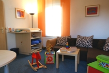 Spielzimmer der Sozialkontaktstelle Wickeltisch, roter Schaukelelch, braunes Sofa mit drei Kissen, kleiner quadratischer Tisch davor und Spielzeug ringsum