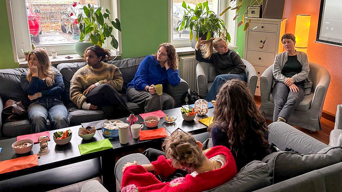 Sieben Personen auf Sofas und Sesseln um einen Coucthtisch mit Gebäckschalen.