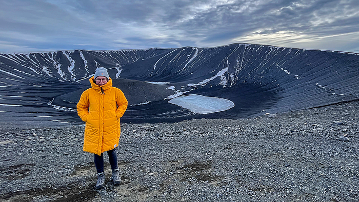 Eine junge Frau steht in Winterkleidung vor einem Vulkankrater. Die Wolkenformation am Himmel wirkt dramatisch.