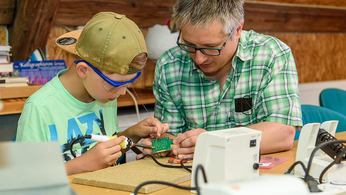 Das Foto zeigt Falko Jahn. Er hält die Platine des Morseapparats in den Händen, auf der zehnjährige David ein Bauteil verlötet.
