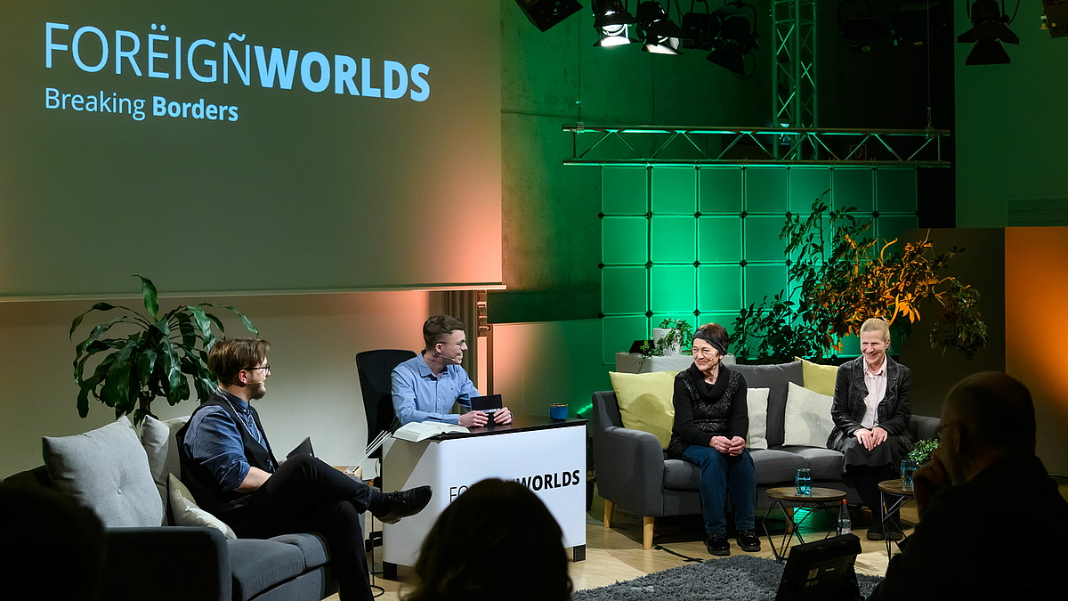 Die beiden Moderatoren wenden ihren Blick zwei weiblichen Personen auf einem Sofa rechts im Bild. Die Stimmung ist heiter.