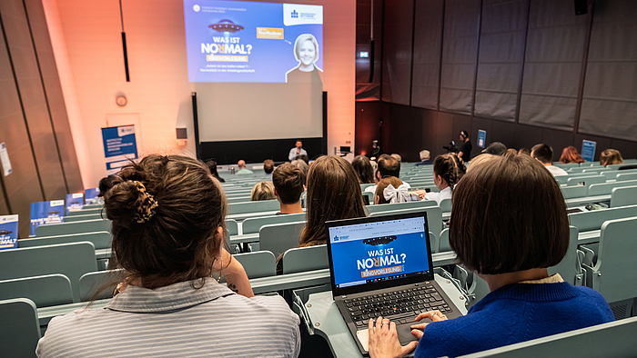 Blick von hinten auf das Publikum in einem Hörsaal. Auf dem Laptop einer Besucherin und projiziert an der Rückwand des Hörsaals ist die Titelfolie zu sehen unter anderem mit dem Text „Was ist normal?' und dem Porträt der heutigen Referentin.