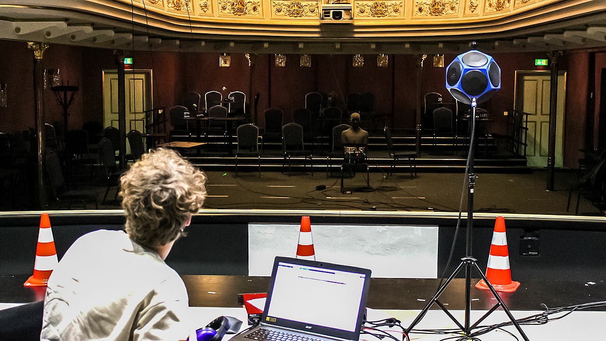 Das Foto blickt in den Zuschauerraum des Theaters mit vereinzelten Stühlen. Am oberen Bildrand ist die reichverzierte Brüstung des ersten Rangs zu sehen. Auf einem der Stühle sitzt der Kunstkopf mit Torso. Ein junger Mann auf der Bühne ist von hinten zu sehen, vor ihm ein aufgeklapptes Laptop. Auf der Bühne befindet sich weiter ein Dodekaeder-Lautsprecher auf einem Stativ mit Laptop sowie am Bühnenrand zum Orchestergraben drei orange-weiße Verkehrsleitkegel.