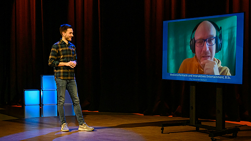 Das Foto zeigt einen jungen Mann und einen großen Monitor vor ienem dunklen Studiohintergrund. Der junge Mann wendet sich einer über den Monitor zugeschalteten Person zu. 