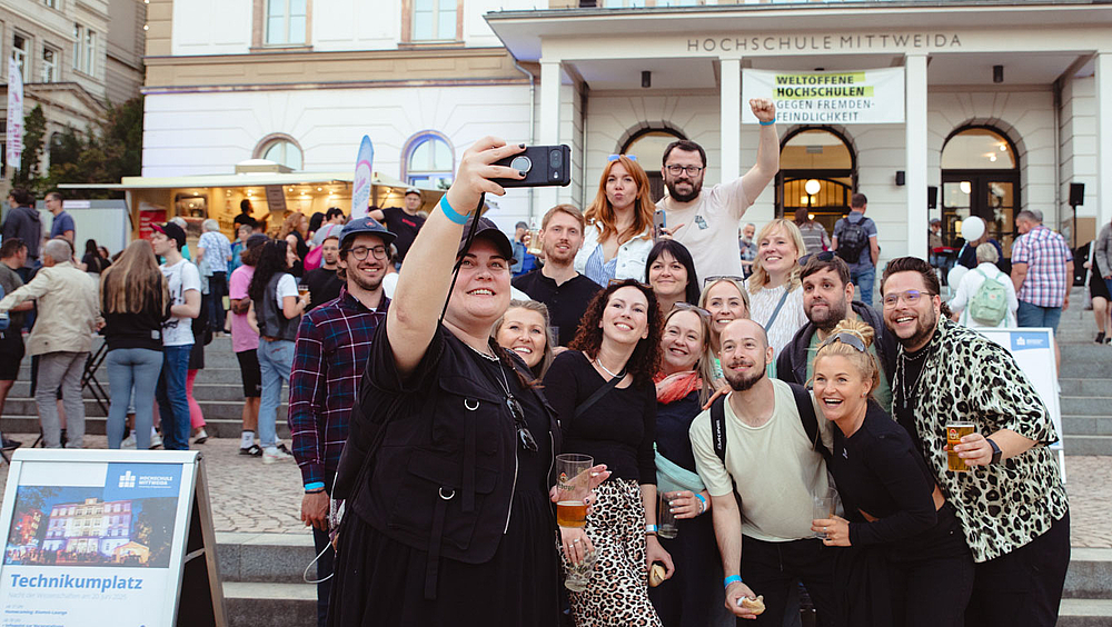 Eine Gruppe von 14 Personen posiert fröhlich für ein Selfie mit dem Hauptgebäude der Hochschule im Hintergrund.