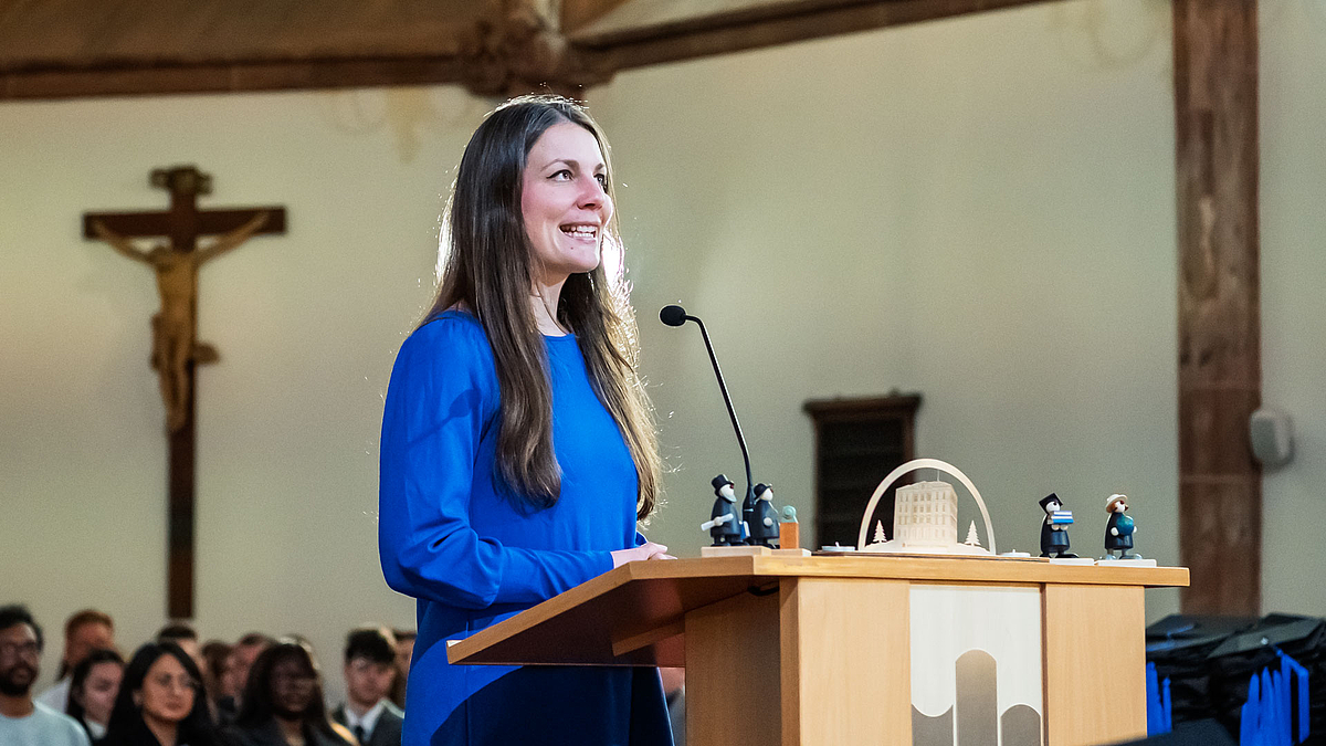 WEibliche Person im blauen Kleid am Rednerpult. Hinrer ihr sind Köpfe von Absolvent:innen zu shen. An der Rückwand des Altarraums hinter ihr hängt ein großes Kruzifix.
