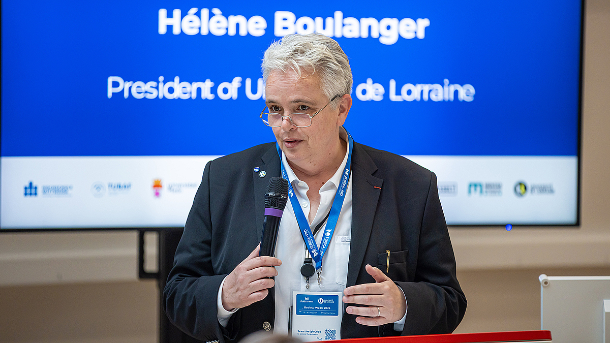 Eine Frau mit kurzen grauen Haaren und Brille steht vor einem Monitor. Darauf ist ihr Name "Hélène Boulanger" eingeblendet und die Logos der EURECA-PRO-Hochschulen abgebildet.