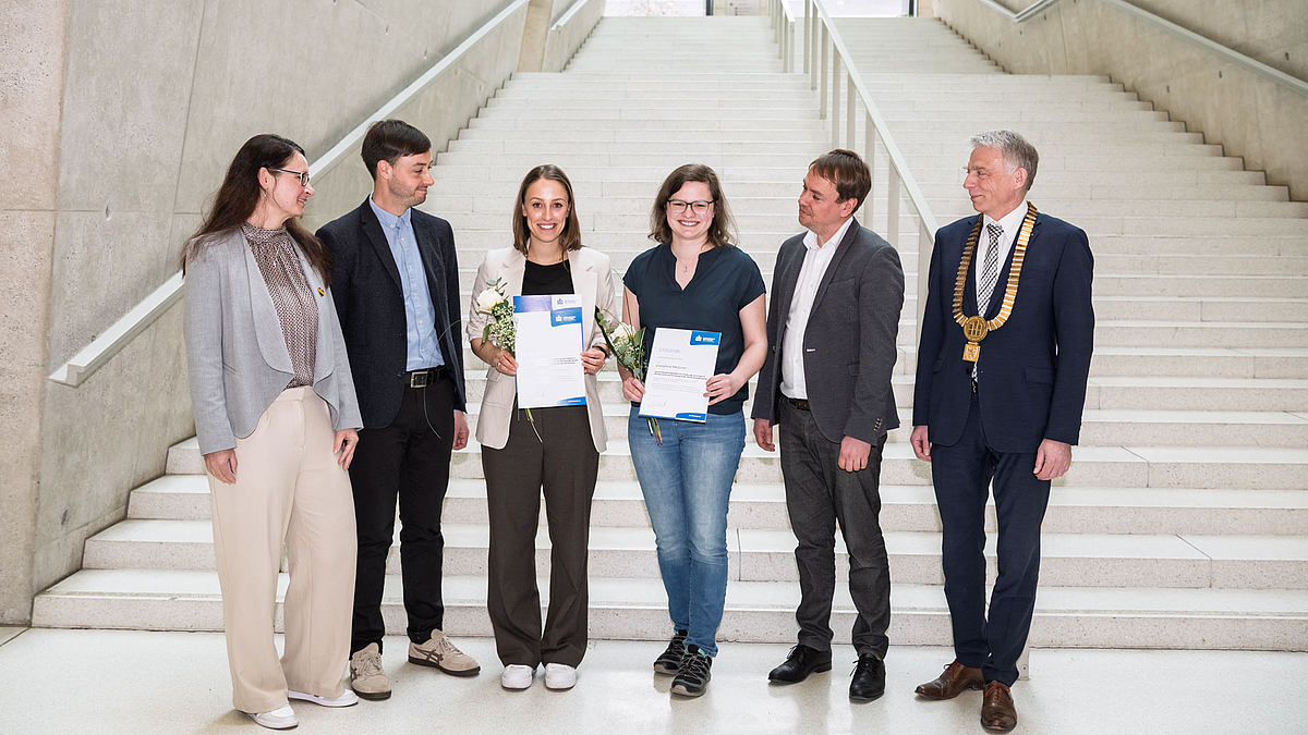 Drei weibliche und drei männliche Personen vor einer großen Treppe. Zwei der weiblichen Personen haben Blumen und Urkunden in der Hand und strahlen. Die anderen Personen schauen zu ihnen.