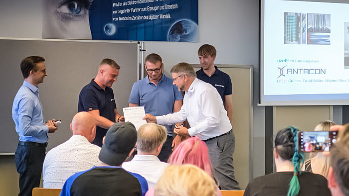 Preis für junge Gründer vom erfahrenen Gründer: Prof. Detlev Müller überreicht beim SAXEED-Gründerstammtisch die Auszeichnung an Hagen Grüttner (2.v.l.), Jan Bretschneider und Johannes Maus von ANTACON. Vor Publikum sind im Bildhintergrund fünf männliche Personen zu sehen. Eine von ihnen bekommt von einer anderen eine Urkunde überreicht (inhalt nicht erkennbar). Auf einem Monitor rechts neben der Personengruppe ist der Schriftzug ANTACON zu erkennen.