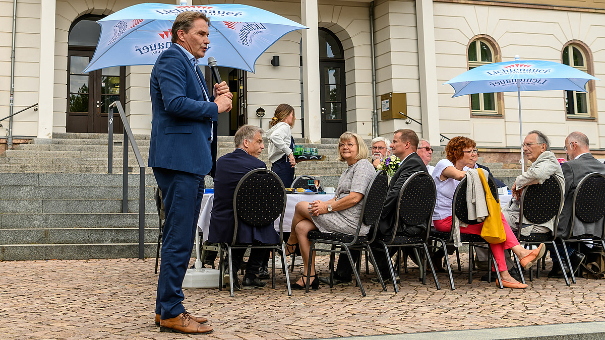 Das Foto zeigt ein männliche Person mit einem Mikrofon in der Hand auf einem Platz stehend. Hinter der Person sitzen Menschen an einem gedeckten Tisch. In Hintergrund ist das Eingansgportal des Hauptgebäudes der Hochschule mit dem Schriftzug Hochschule Mittweida.