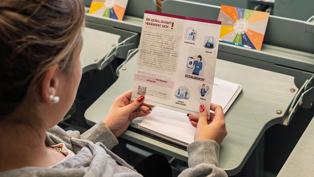 Person von hinten im Anschnitt, die auf einen Flyer mit der Überschrift "Die Gesellschaft verändert sich" schaut.