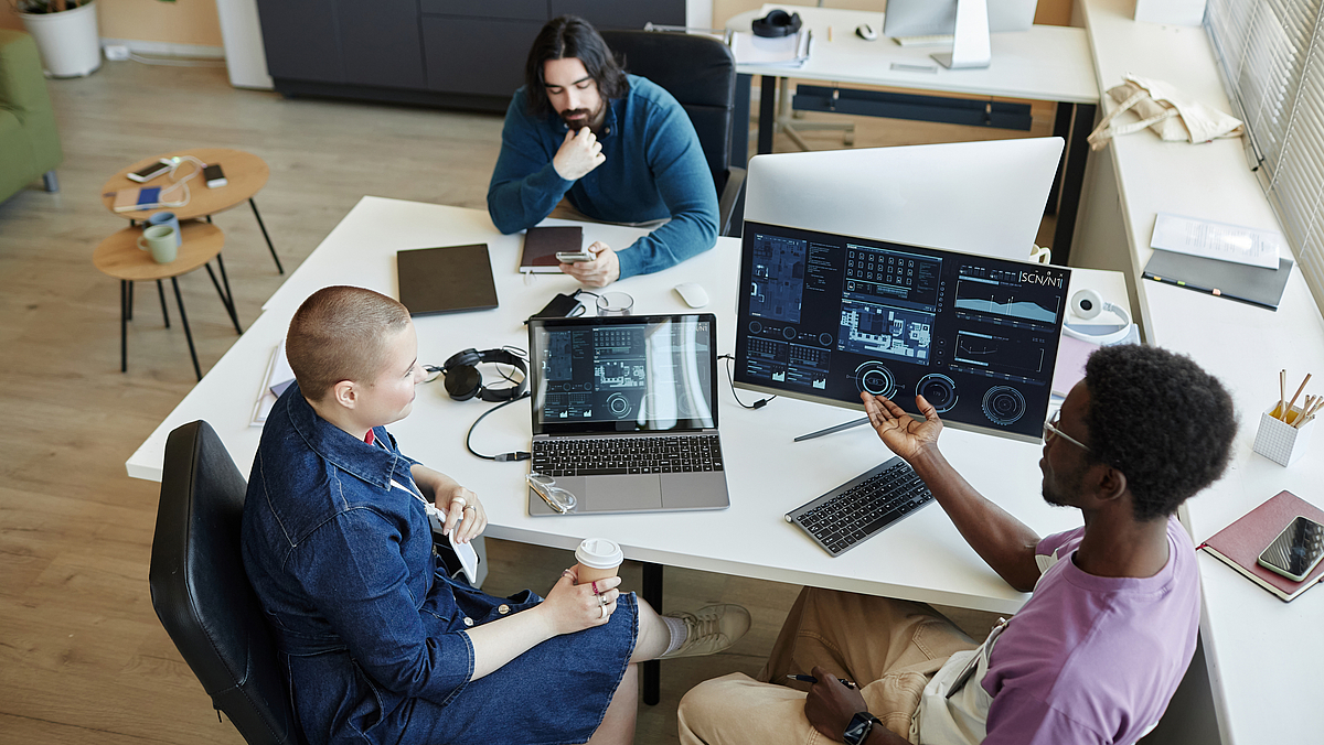 Eine Frau und zwei Männer sitzen an beiden langen Seiten eines großen Schreibtischs, auf dem ein Laptop und ein Monitor mit Datenvisualisierungen stehen und besprechen sich.