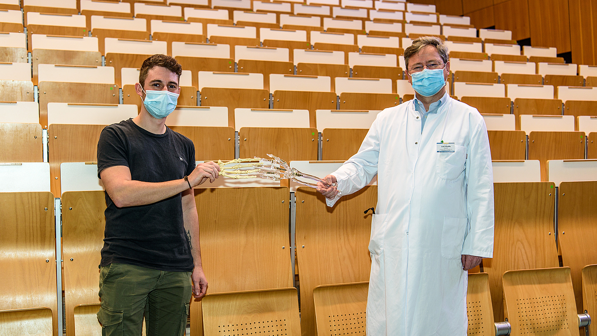 Das Foto zeigt zwei Personen mit medizinischer Maske vor den unbesetzten ansteigenden Stuhlreihen eines Hörsaals. Sie blicken in die Kamera Die Person rechts trägt einen weißen Arztkittel und hält das Modell eines Unterarmknochens mit Hand aus Plexiglas der Person links entgegen, die ein ebensolches aber nicht durchsichtiges Modell hält. Die beiden Skeletthänden berührten sich wie für Handschlag.