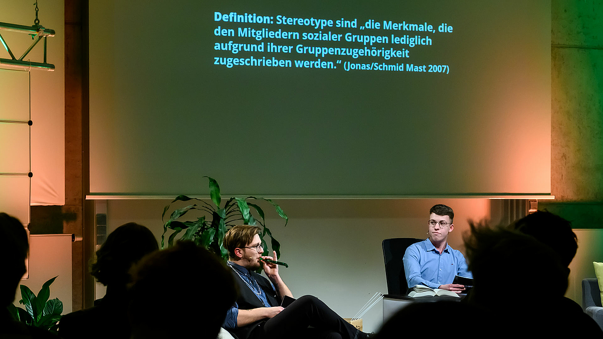 Zwei junge männliche Personen sitzen unterhalb einer Leinwand, auf die eine Definition projiziert ist: „Stereotype sind die Merkmale, die den Mitgliedern sozialer Gruppen lediglich aufgrund ihrer Gruppenzugehörigkeit zugeschrieben werden.“