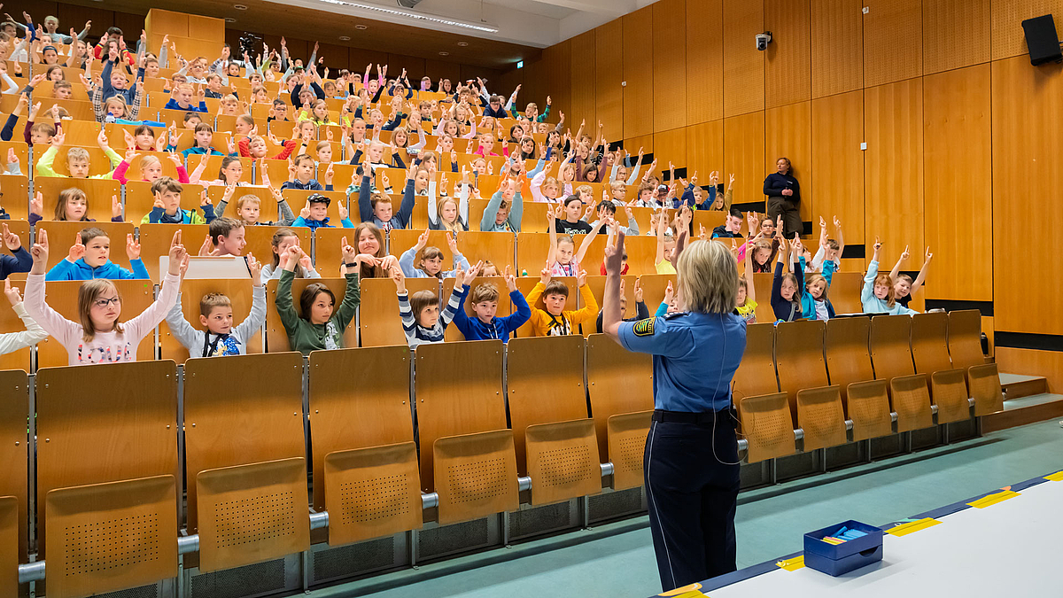 Polizistin in Uniform vor mit Kindern vollbesetztem Hörsaal. Alle heben die Hände zum leisen Klastchen mit je zwei Fingern.