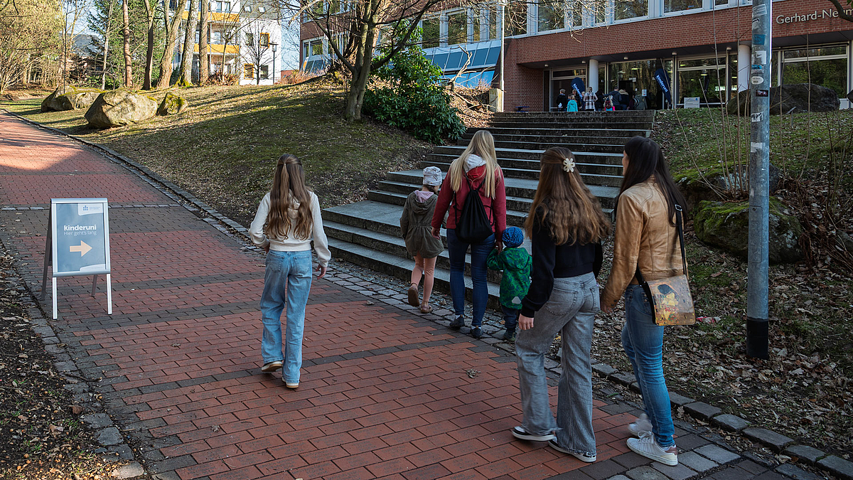 Kinder und Erwachsene auf dem Weg zum Hörsaalgebäude. Auf dem Weg steht ein Kinderuni-Aufsteller.