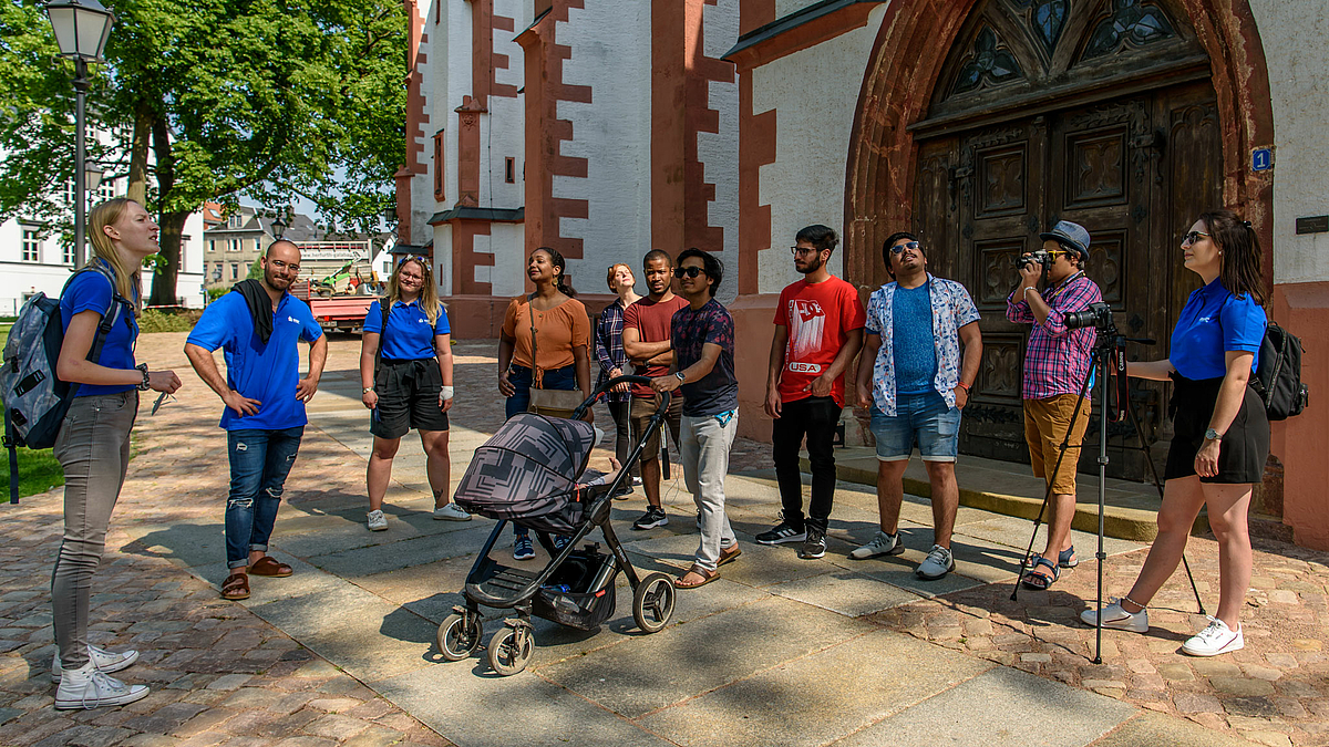 Das Foto zeigt eine Gruppe von elf jungen Menschen vor dem Seitenportal der Evangelischen Stadtkirche Mittweida. Einige blicken nach oben. Vier von ihnen tragen blaue Hochschul-Shirts. Sie sind die Guides der internationalen Studentengruppe auf ihrer Besichtigungstour durch Mittweida.