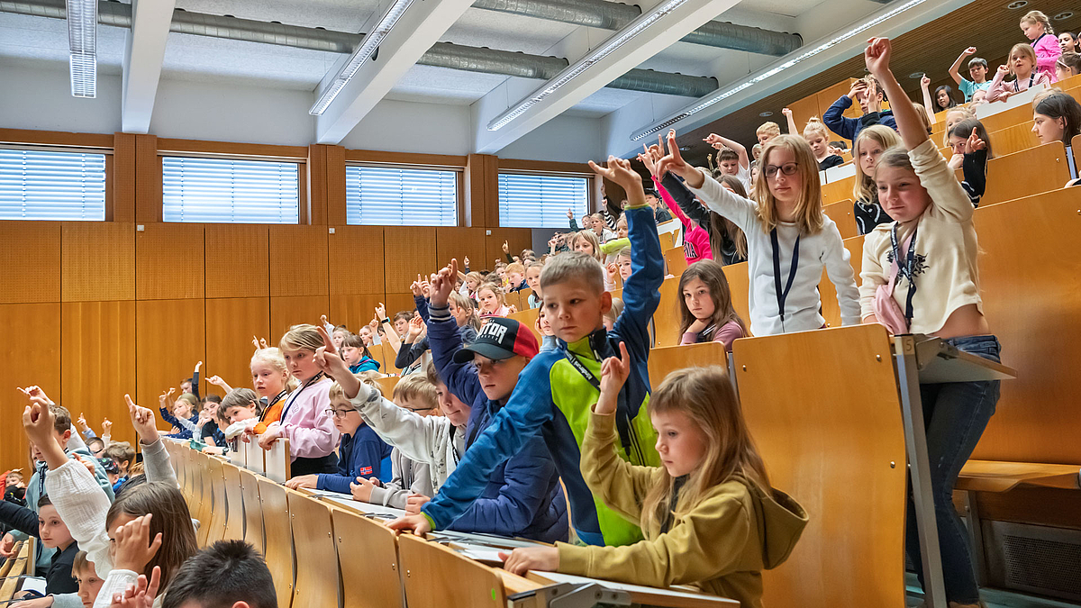Viele Kinder sitzen in einem Hörsaal und heben die Hände.
