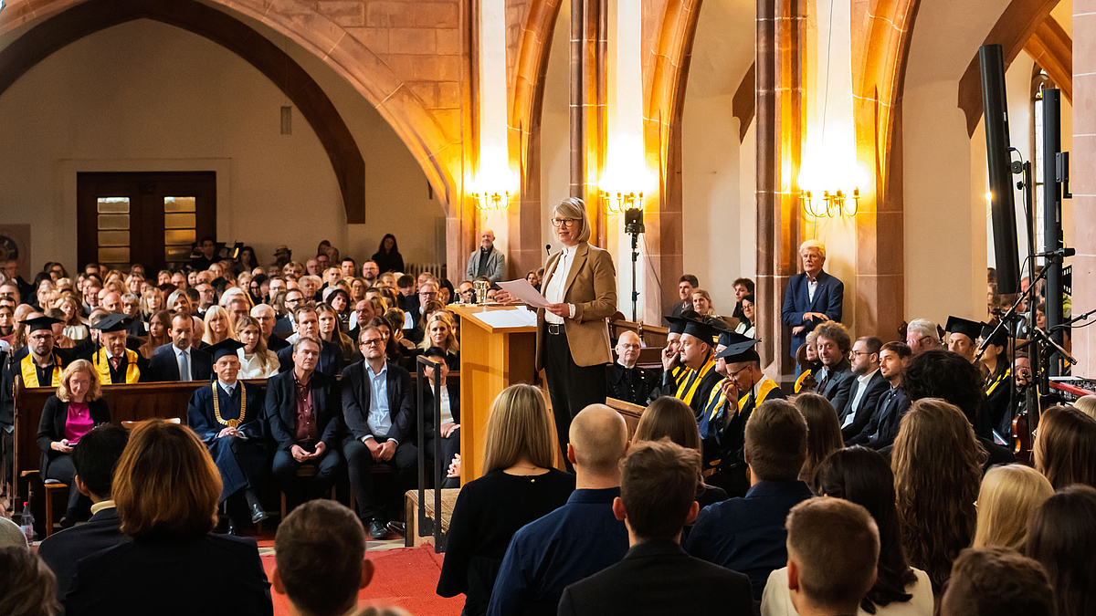 Blick vom Altarraum auf eine weibliche Person am Rednerpult wendet sich den Absolvent:innen hinter sich zu.