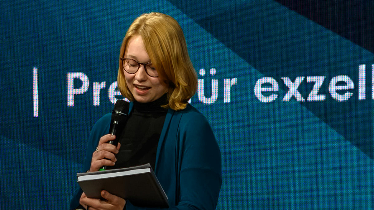 Das Foto zeigt eine junge Frau mit Mikrofon in der Hand, den Blick auf ihr Manuskript gerichtet. Hinter ihr auf dem Display ist unvollständig der Titel "Preis für exzellente ..." zu erkennen. l