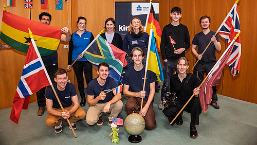 Mission completed: Das Kinderuni-Team der Hochschule macht Globalisierung und andere spannende Themen erlebbar. Zehnhn Personen stehen und knien im Hörsaal, einige halten verschiedene Nationalflaggen hoch und lächeln. Hinter der Gruppe ist ein Kinderuni-Rollup.