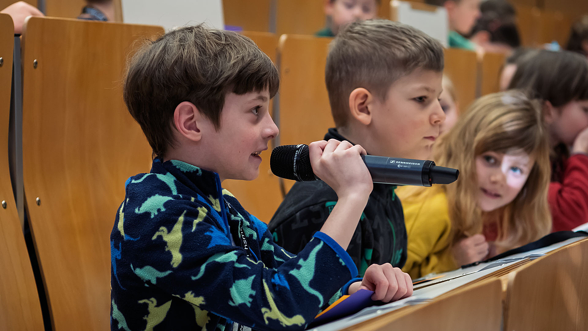 : Ein Junge in einem Hörsaal hält ein Mikrofon und spricht hinein. Ein Mädchen daneben lehnt sich vor und schaut ihn freundlich an.